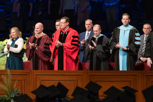 Elder Clark G. Gilbert (center), and BYU–Idaho President Alvin F. Meredith III and his wife, Jennifer (left), applaud as graduates enter the BYU–Idaho Center for commencement on Thursday, December 19, 2024. Photo by Reilly Cook, BYU–Idaho. Elder Gilbert’s call to the Quorum of the Twelve Apostles have led to questions about his commitment to BYU academic freedom.
