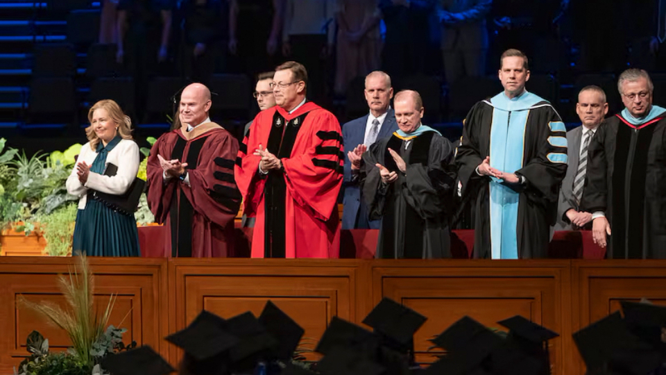 Elder Clark G. Gilbert (center), and BYU–Idaho President Alvin F. Meredith III and his wife, Jennifer (left), applaud as graduates enter the BYU–Idaho Center for commencement on Thursday, December 19, 2024. Photo by Reilly Cook, BYU–Idaho. Elder Gilbert’s call to the Quorum of the Twelve Apostles have led to questions about his commitment to BYU academic freedom.