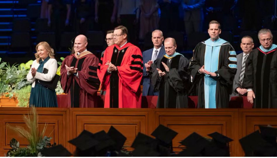 Elder Clark G. Gilbert (center), and BYU–Idaho President Alvin F. Meredith III and his wife, Jennifer (left), applaud as graduates enter the BYU–Idaho Center for commencement on Thursday, December 19, 2024. Photo by Reilly Cook, BYU–Idaho. Elder Gilbert’s call to the Quorum of the Twelve Apostles have led to questions about his commitment to BYU academic freedom.