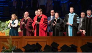Elder Clark G. Gilbert (center), and BYU–Idaho President Alvin F. Meredith III and his wife, Jennifer (left), applaud as graduates enter the BYU–Idaho Center for commencement on Thursday, December 19, 2024. Photo by Reilly Cook, BYU–Idaho. Elder Gilbert’s call to the Quorum of the Twelve Apostles have led to questions about his commitment to BYU academic freedom.
