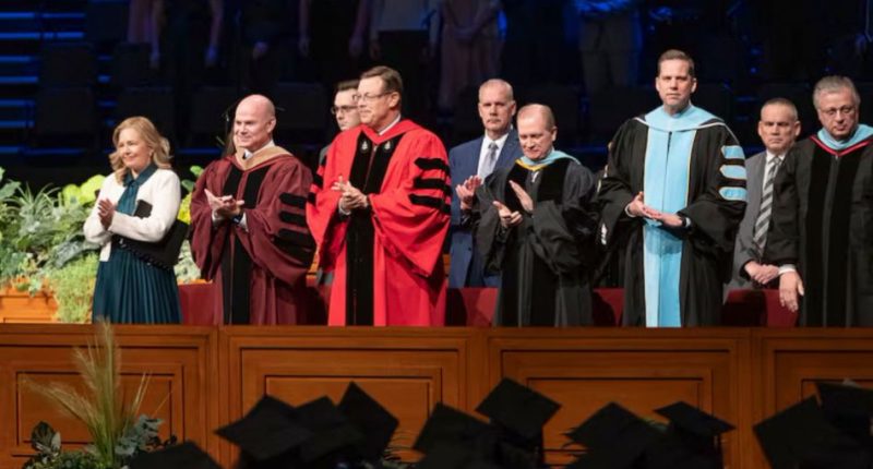 Elder Clark G. Gilbert (center), and BYU–Idaho President Alvin F. Meredith III and his wife, Jennifer (left), applaud as graduates enter the BYU–Idaho Center for commencement on Thursday, December 19, 2024. Photo by Reilly Cook, BYU–Idaho. Elder Gilbert’s call to the Quorum of the Twelve Apostles have led to questions about his commitment to BYU academic freedom.