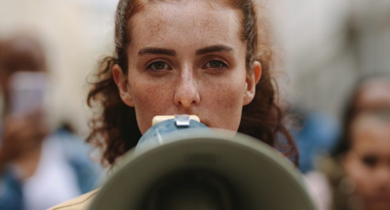 Female activist protesting with megaphone during a strike with group of demonstrator in background. Woman protesting in the city.